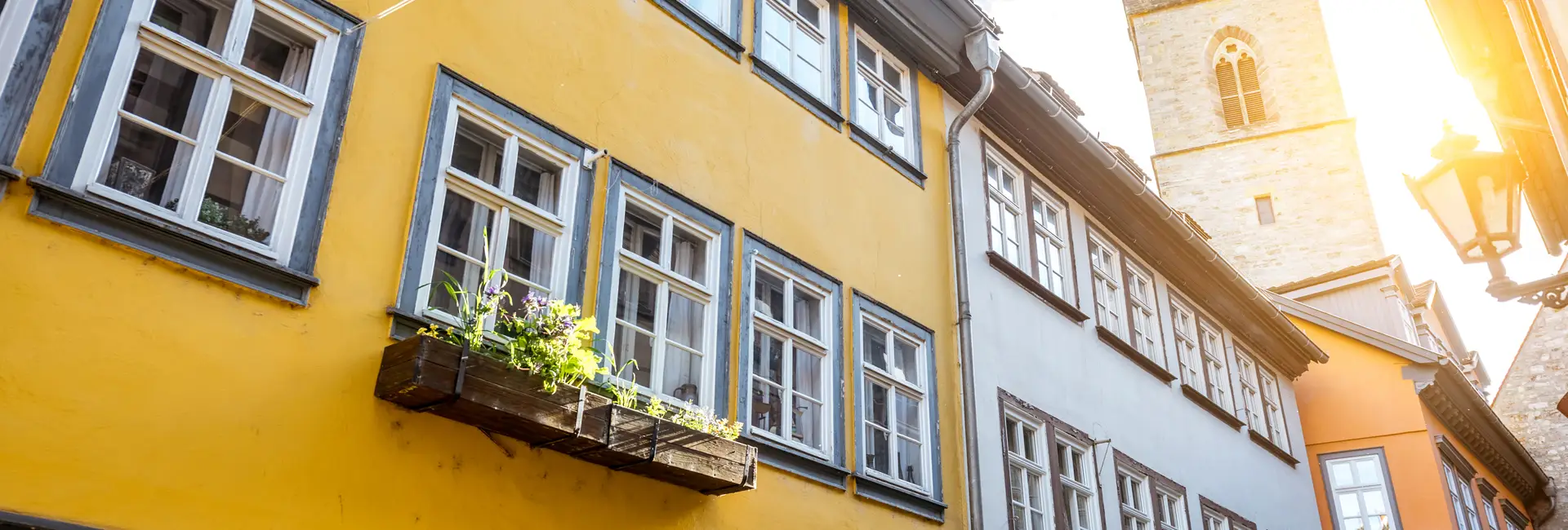 Historische Gebäude mit gelben und blauen Fassaden, mehreren Fenstern und einem Blumenkasten unter einem klaren Himmel, während im Hintergrund Sonnenlicht über einen Glockenturm strömt.