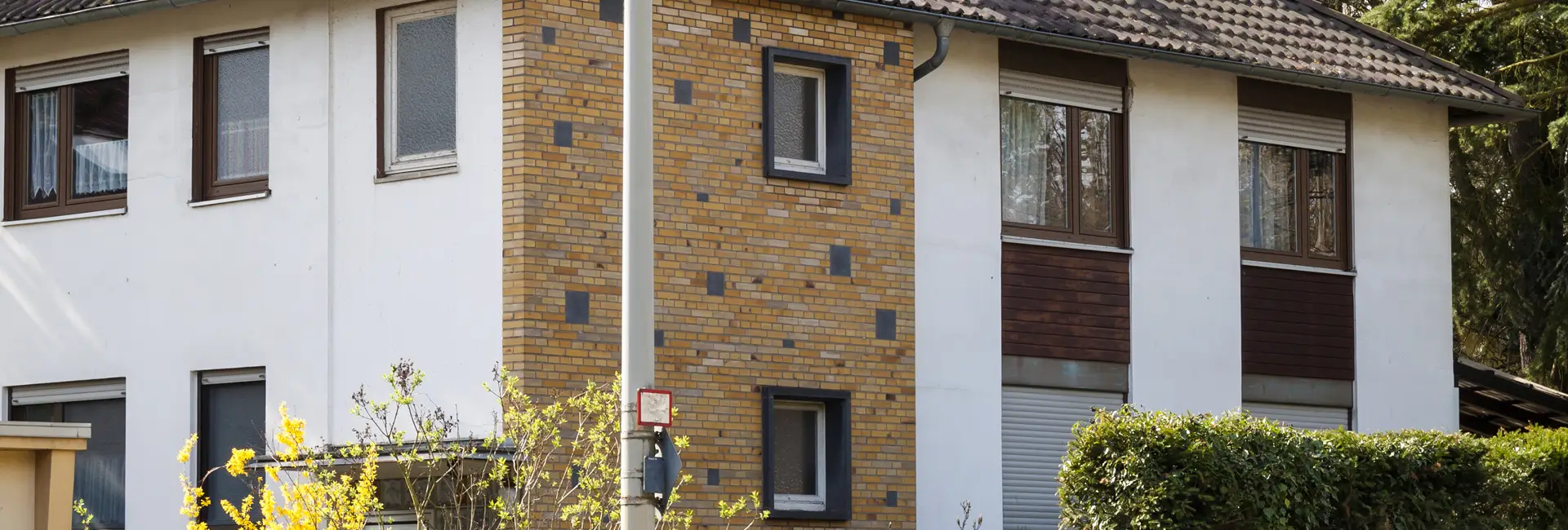 Zweistöckiges Haus mit braunen Ziegeln und weißer Außenverkleidung, mehreren Fenstern, einem Ziegeldach und umgebendem Grün.