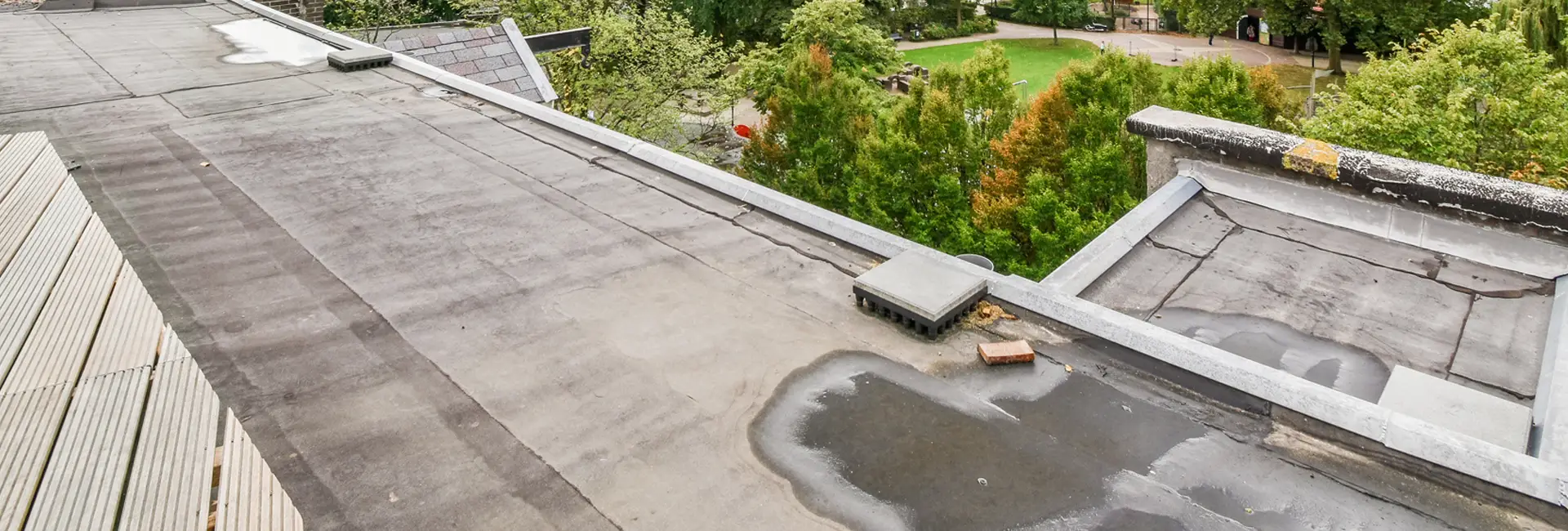 Flaches Dach mit Betonoberfläche, einer kleinen Pfütze und einem Lüftungsschacht. Im Hintergrund sind Bäume und ein grasbewachsener Park zu sehen.