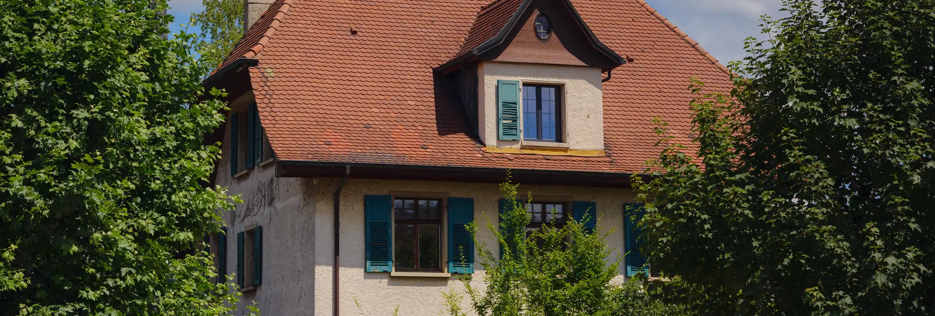 Ein zweistöckiges Haus mit einem steilen, roten Ziegeldach und grünen Fensterläden, umgeben von grünen Bäumen unter einem blauen Himmel.