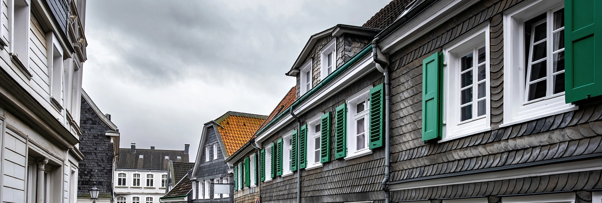 Schmale Straße gesäumt von traditionellen europäischen Häusern mit grauen Schieferfassaden und grünen Fensterläden unter einem bewölkten Himmel.